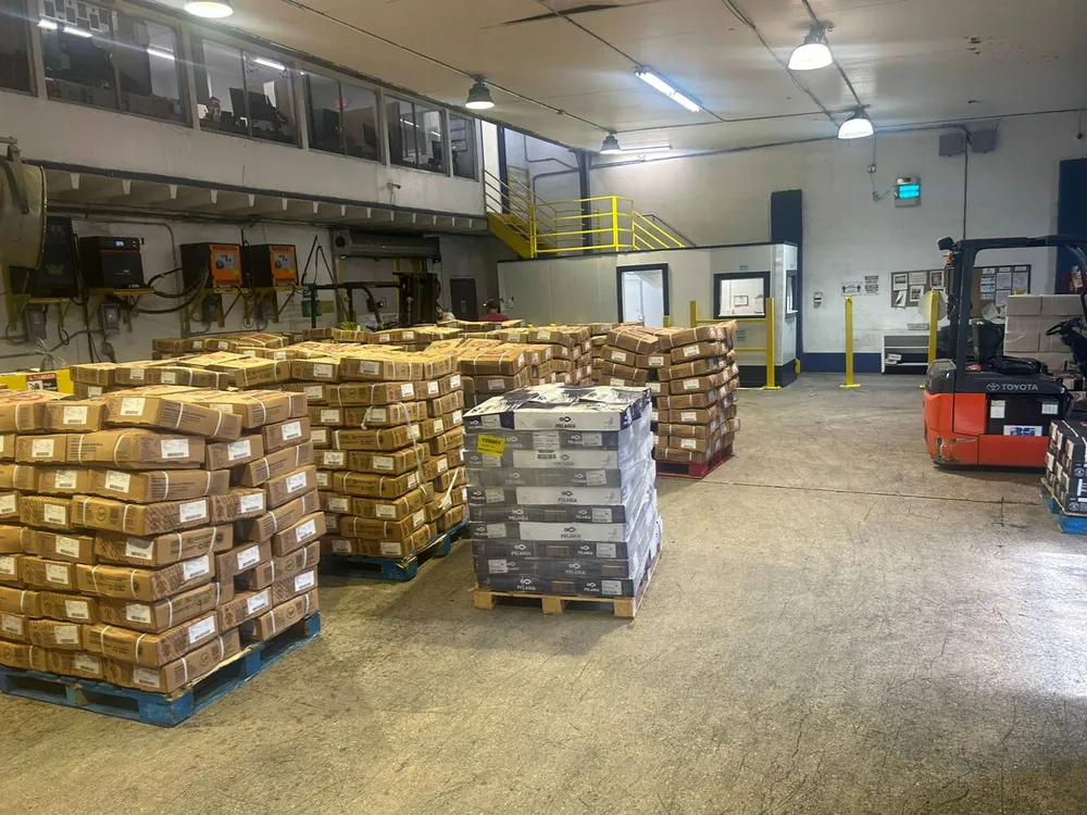 Pallets of cardboard cases on a warehouse floor, with a forklift, yellow stairs and a raised office level behind