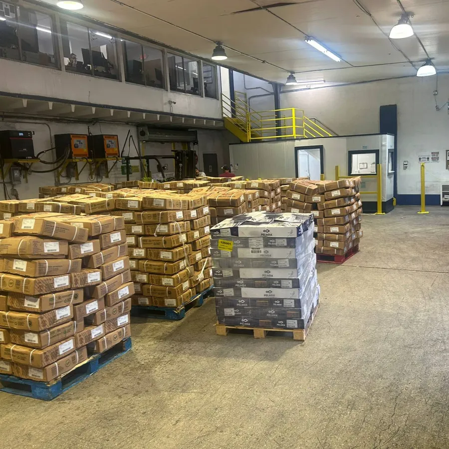 Pallets of stacked cardboard cases on a warehouse floor, with a forklift and a raised office level behind
