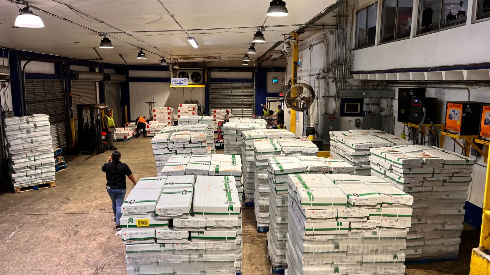 Warehouse floor with rows of palletized, shrink-wrapped boxes of product while staff and a forklift work near the loading docks